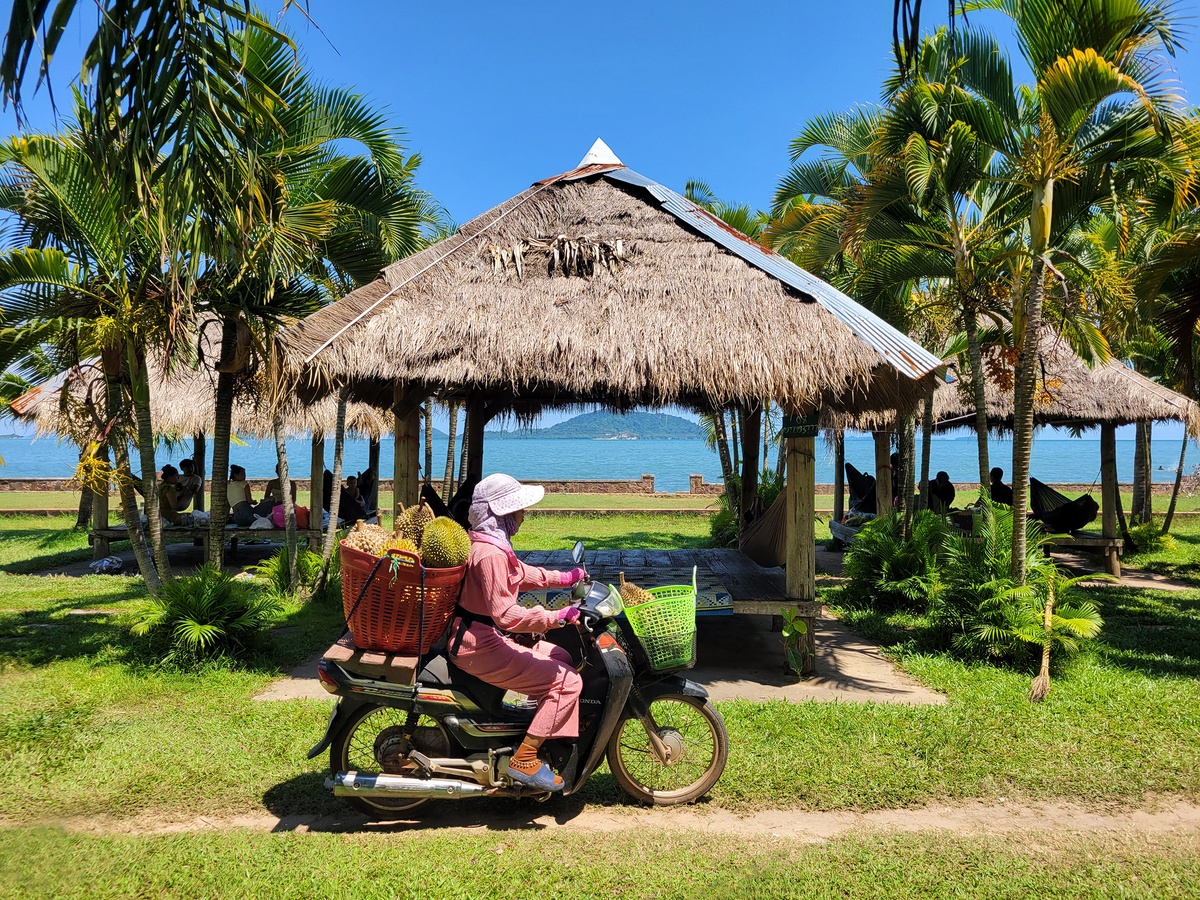 Traditional bungalow in Kep, Cambodia, with a woman passing by on a motorbike carrying durians